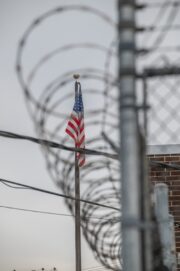 American flag behind a chain-link fence topped with barbed wire, symbolizing immigration and border security.