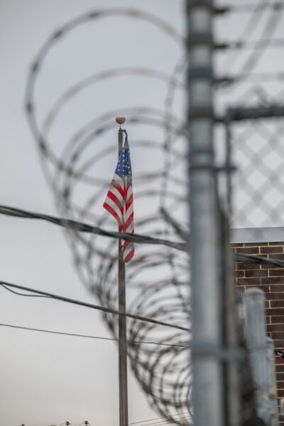 American flag behind a chain-link fence topped with barbed wire, symbolizing immigration and border security.