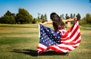 Child sitting on a grassy field outdoors while holding a large American flag spread around the body.