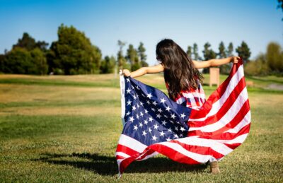 Child sitting on a grassy field outdoors while holding a large American flag spread around the body.