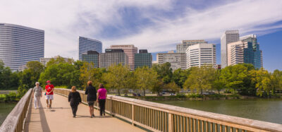 People walking across a pedestrian bridge over water with trees and the Arlington County skyline visible in the background.